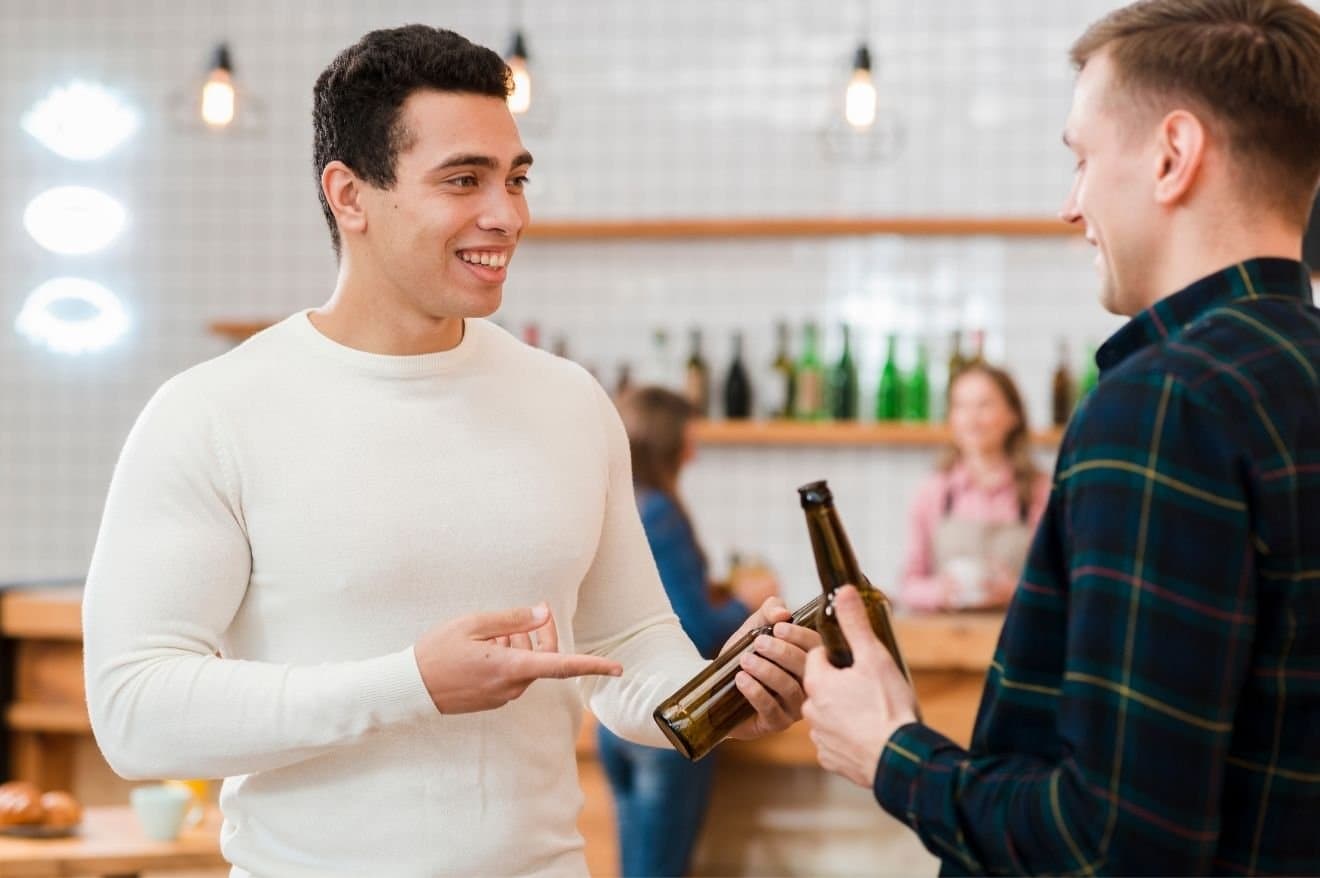 How to Tell if an Alcohol Bottle Has Been Opened and Resealed 2 Two men engaged in conversation at a bar, with drinks on the table and a lively atmosphere in the background.